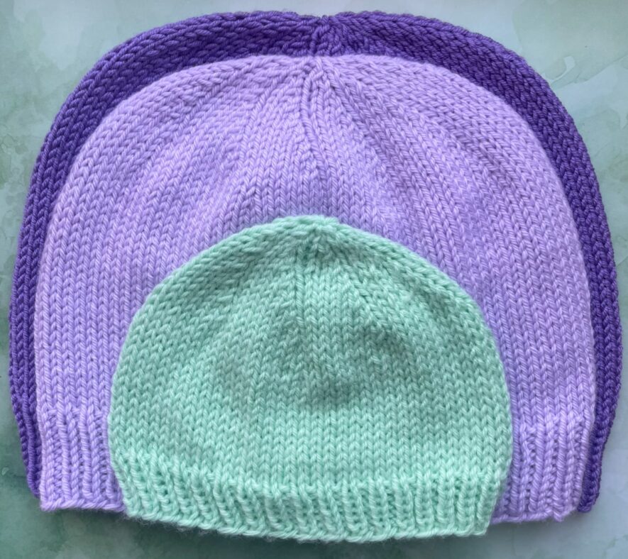 Green newborn DK hat on top of child pale purple DK hat on top of adult dark purple DK hat
