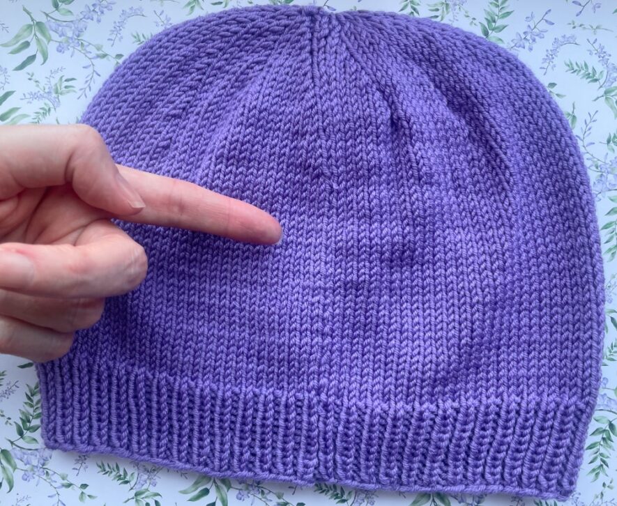 Hand pointing to mattress stitch seam down back of DK beanie knit with straight needles