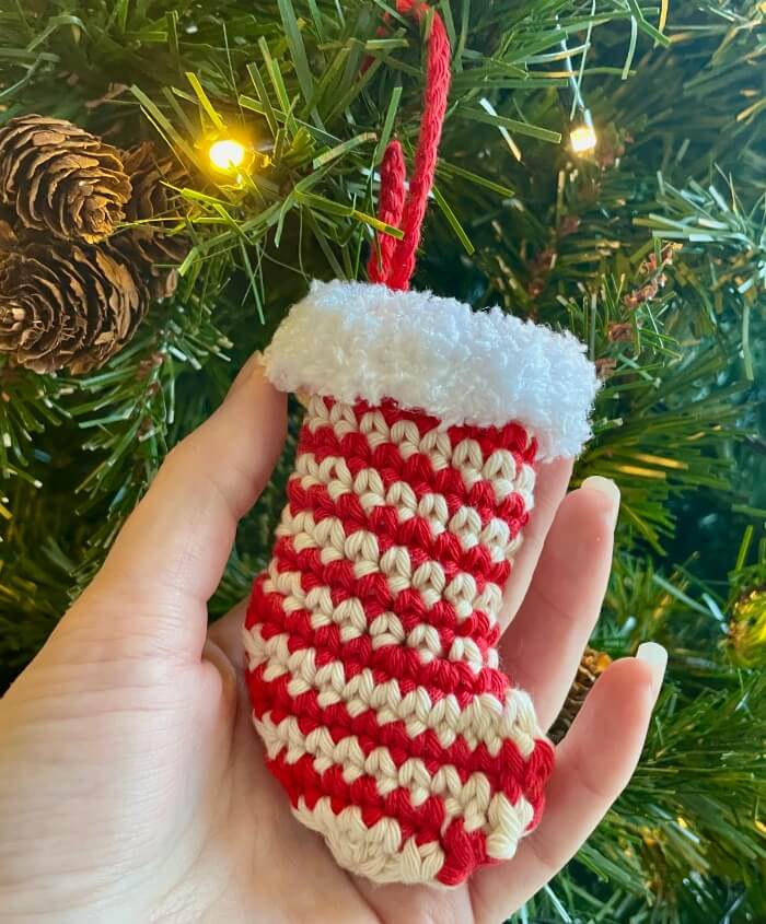 Close up of hand holding red and white striped crochet mini stocking with fluffy white cuff hanging on Christmas tree