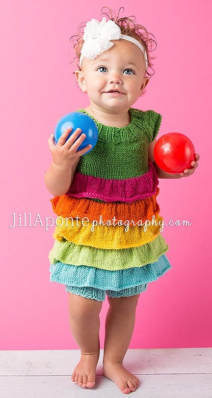 Toddler wearing rainbow ruffle dress free knitting pattern by Claire Gentry and holding toy balls